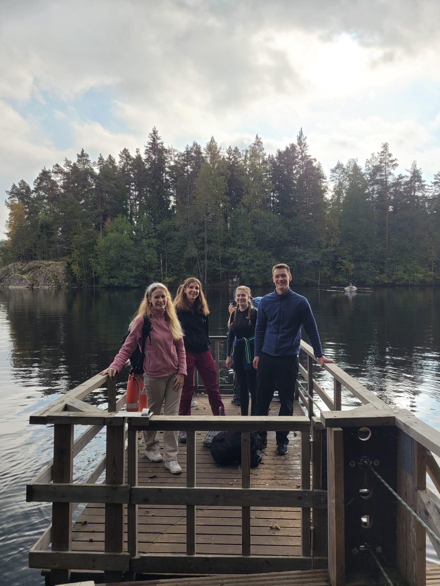 Four hikers smiling together on a wooden platform beside a forest lake